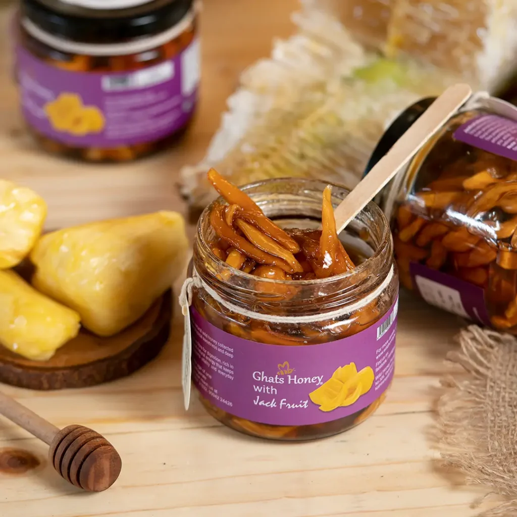 Ghats Honey with Jackfruit – ripe jackfruit slices soaked in pure paalaipoo honey and stored in a glass jar with fresh jackfruit and honeycomb on a wooden table.